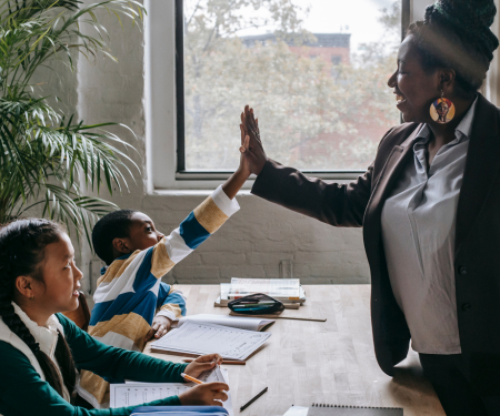 Black woman teacher high fiving a black male student at his desk