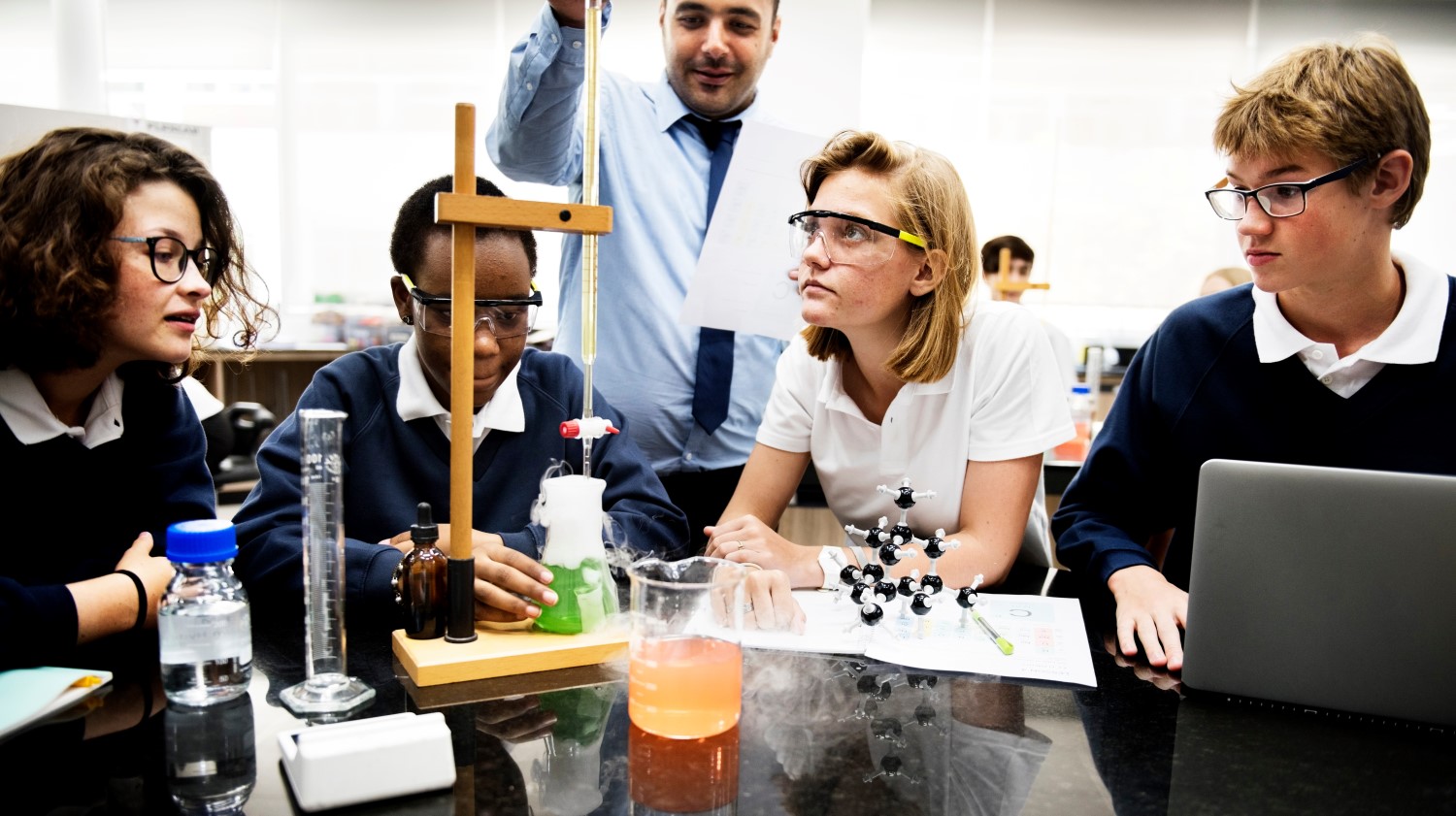 Group of science students around an experiment with their teacher overlooking