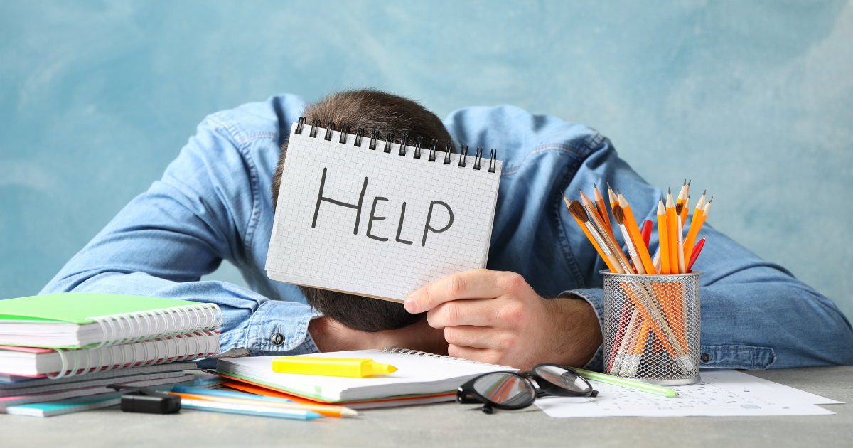 Male teacher at a desk with notebooks and pencils, holding up a sign that reads 'help'