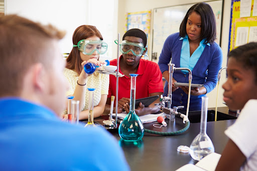 A group of high school students in a lab, surrounded by chemistry equipment with their teacher observing