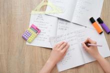 A hand writing math equations in a notebook with a calculator and highlighters on the table