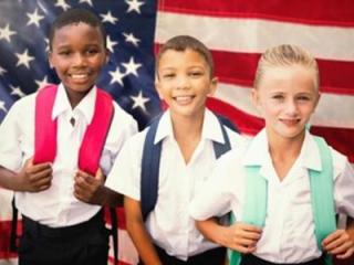 Three diverse children wearing backpacks and standing in front of an American flag