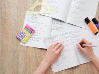 A hand writing math equations in a notebook with a calculator and highlighters on the table